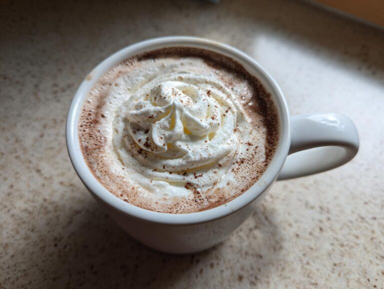 Close-up of a mug filled with rich white hot chocolate topped with a swirl of whipped cream and a dusting of cocoa.