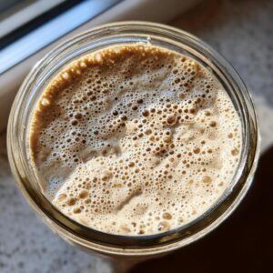 Overhead view of an active sourdough starter in a glass jar, showing lots of small, active bubbles on the surface.