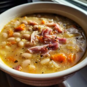 Close-up of a warm bowl of navy bean soup, featuring white beans, chunks of ham, and orange carrots, seasoned with pepper.