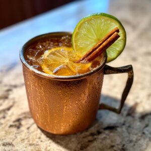 A close-up of a frosty apple cider moscow mule garnished with a lime wheel and cinnamon sticks in a copper mug.