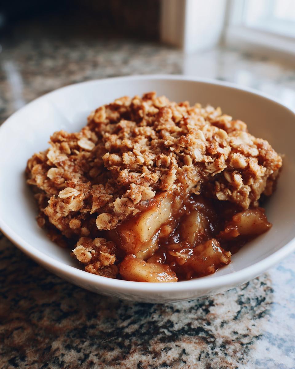 Close-up of a single serving of the best apple crisp recipe in a white bowl, showing warm, spiced apples beneath a crunchy oat topping.