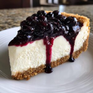 A perfect slice of creamy blueberry cheesecake with a thick graham cracker crust and rich blueberry topping dripping down the side.