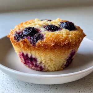 A single, perfectly baked blueberry cottage cheese muffin with a golden top and visible blueberries, resting on a small white plate.