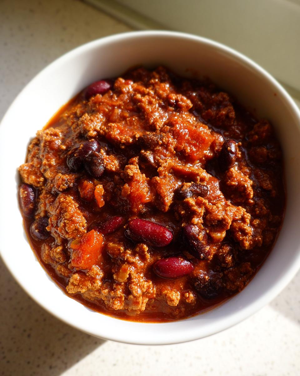 Close-up overhead view of a white bowl filled with thick, rich high protein chili featuring ground meat and dark red kidney beans.