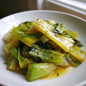 A close-up serving of tender, glossy braised leeks coated in a savory, herb-flecked sauce.
