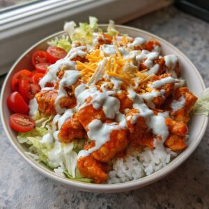 A close-up of a fully assembled buffalo chicken bowl featuring rice, lettuce, orange buffalo chicken pieces, ranch dressing, and shredded cheese.