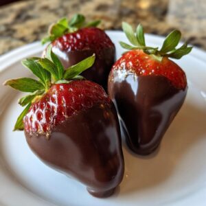Close-up of three glossy chocolate covered strawberries, lightly dusted with gold shimmer, resting on a white plate.