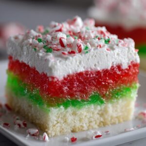 Close-up of a layered Christmas poke cake slice featuring white cake, red and green gelatin, whipped topping, and crushed peppermint.