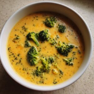 Close-up overhead shot of a white bowl filled with creamy, orange-yellow broccoli cheddar soup featuring bright green broccoli florets.