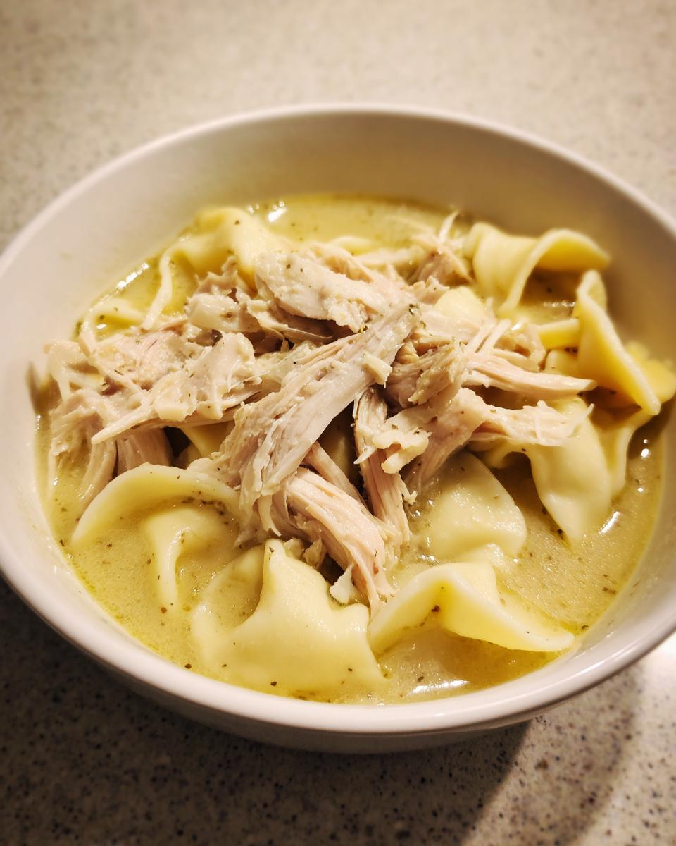 A close-up of a white bowl filled with creamy chicken and dumplings, topped generously with shredded seasoned chicken.