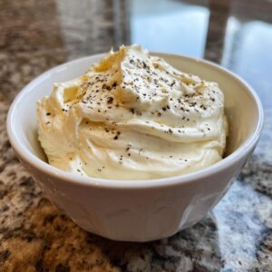 A close-up of creamy horseradish sauce, swirled high in a white bowl and sprinkled with coarse black pepper.