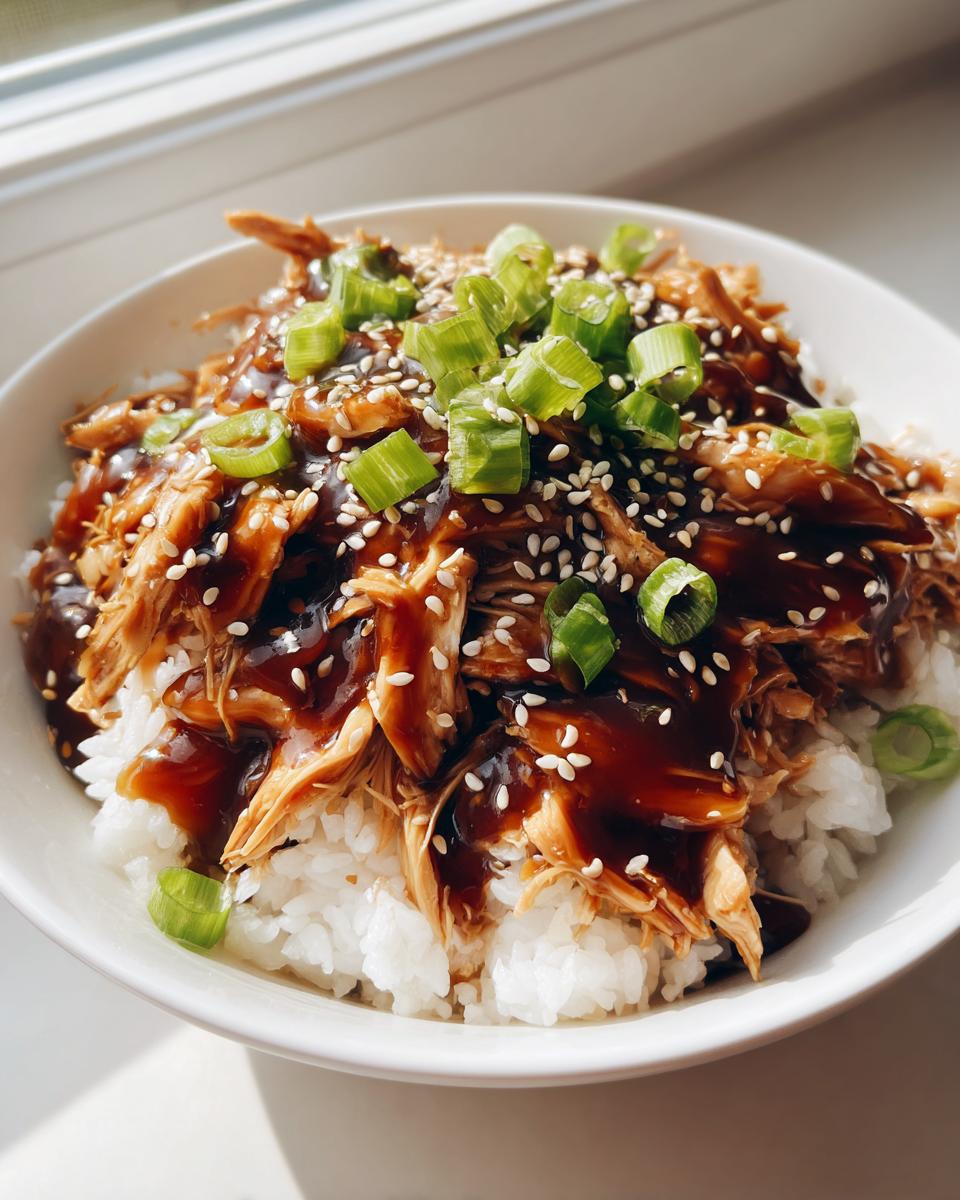 A close-up of a bowl filled with white rice topped with shredded crockpot teriyaki chicken, glossy sauce, sesame seeds, and green onions.