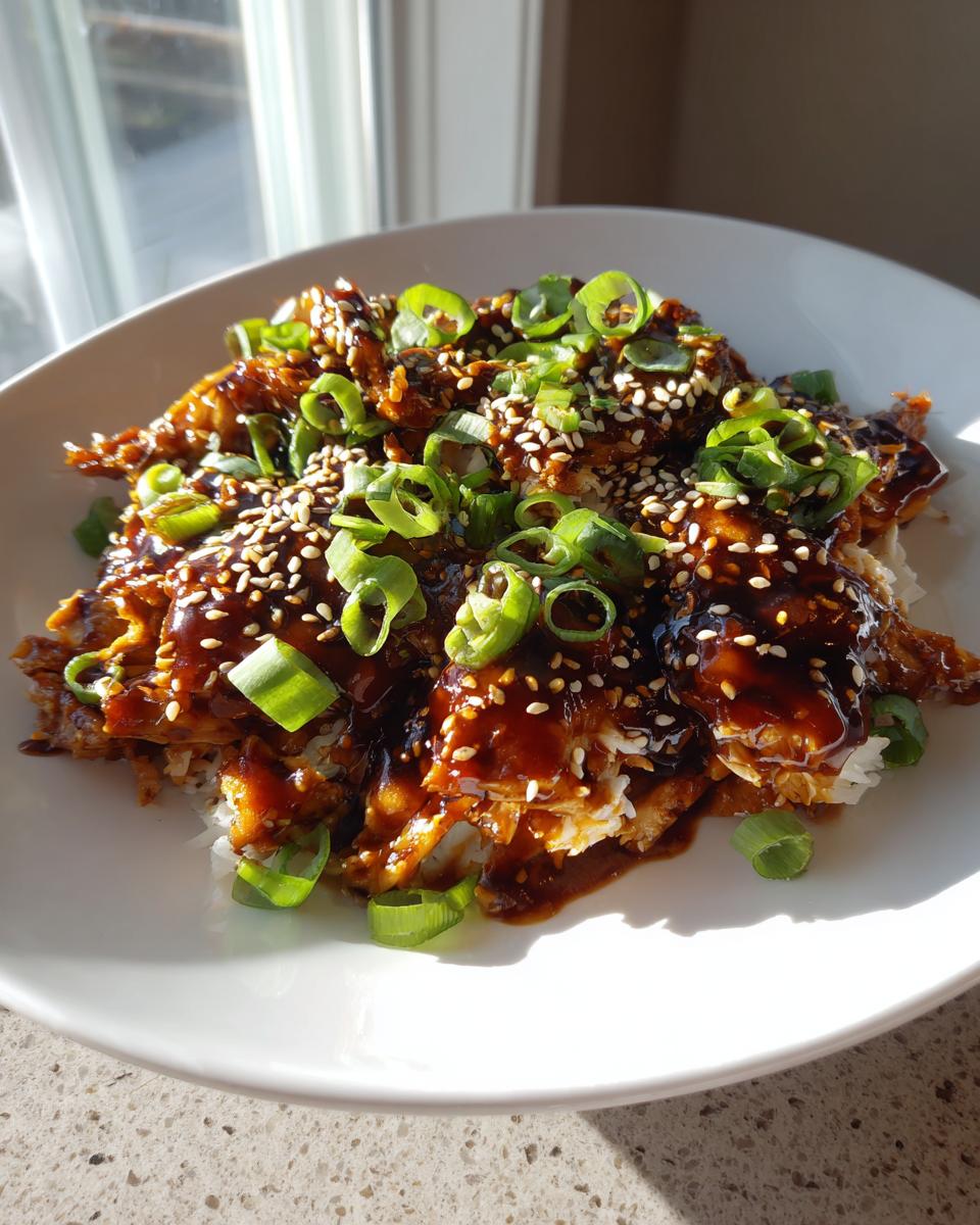 Shredded crockpot teriyaki chicken coated in thick sauce, topped with green onions and sesame seeds, served over white rice.
