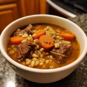 Close-up of a white bowl filled with rich beef barley soup, featuring chunks of beef, bright carrots, and cooked barley.