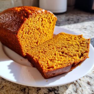A loaf of rich, orange pumpkin bread with one slice cut and resting against the main loaf on a white plate.