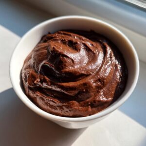 A close-up of thick, glossy, dark brown chocolate dip swirled in a small white bowl, catching bright sunlight.