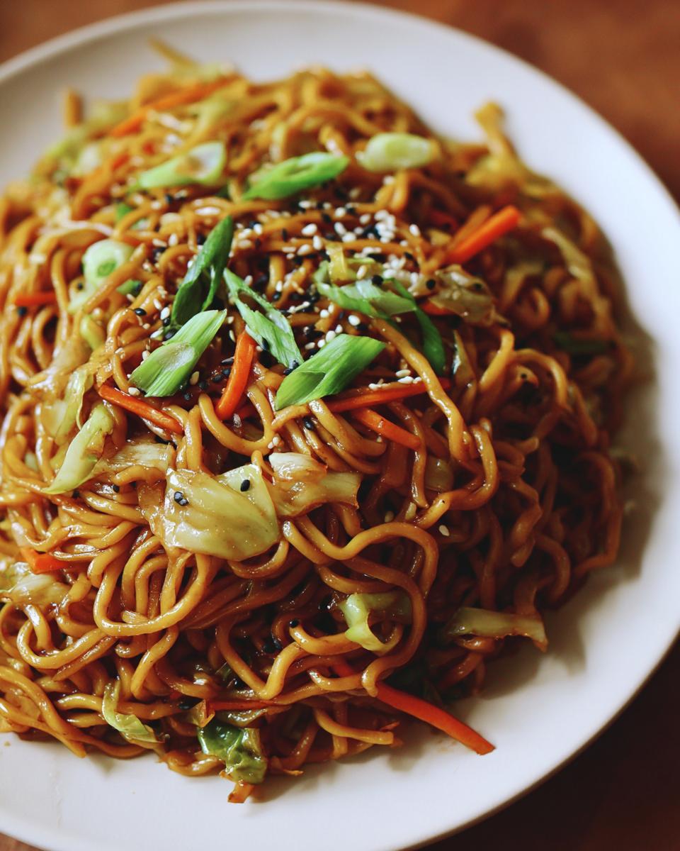 A close-up of a plate piled high with savory fried noodles mixed with cabbage and carrots, topped with green onions and sesame seeds.