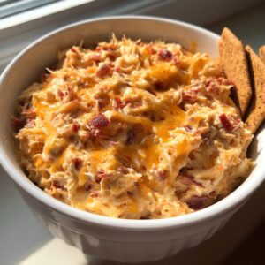 A close-up of warm, cheesy Reuben dip topped with melted cheddar and bits of corned beef in a white bowl, served with crackers.